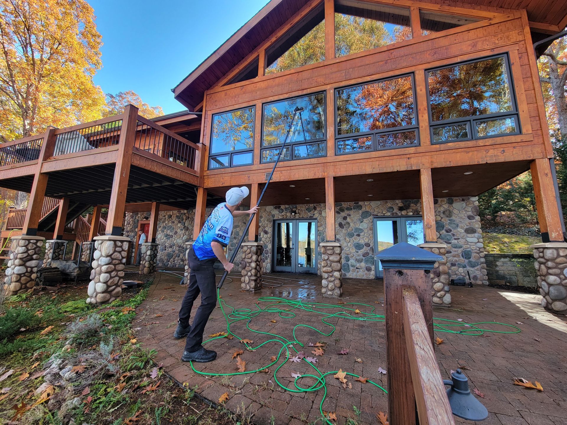 Person washing windows of a large wood and stone house with water hose. Autumn foliage in background.