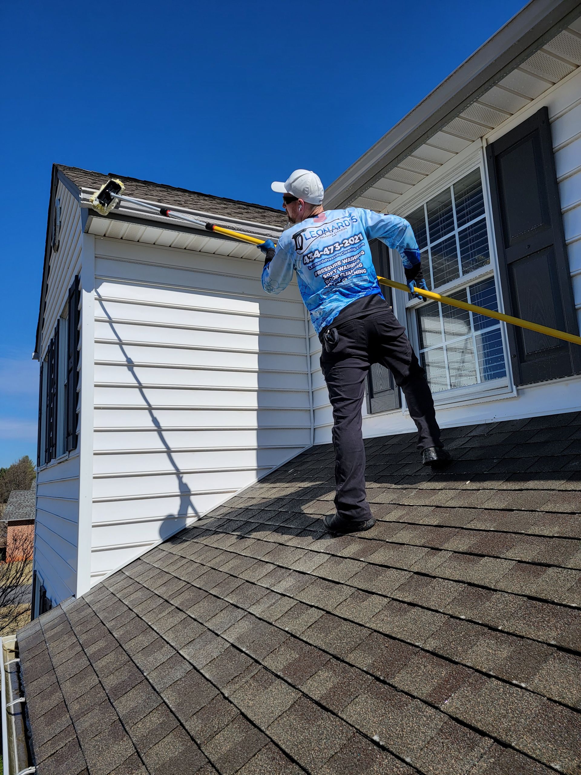 Man on roof cleaning gutters with a pole; white house, blue sky.