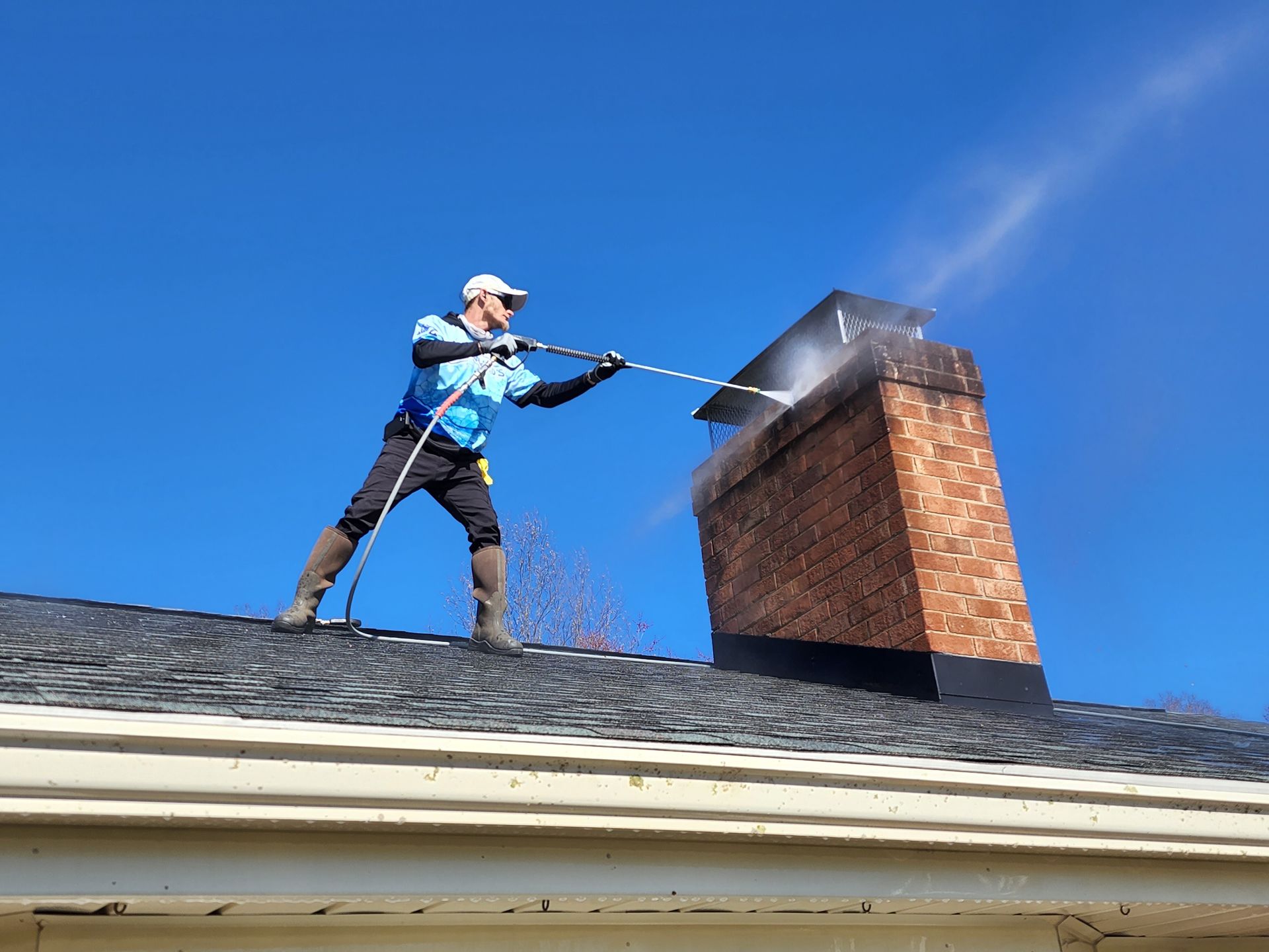 Man power washing a brick chimney on a roof with blue sky in the background.