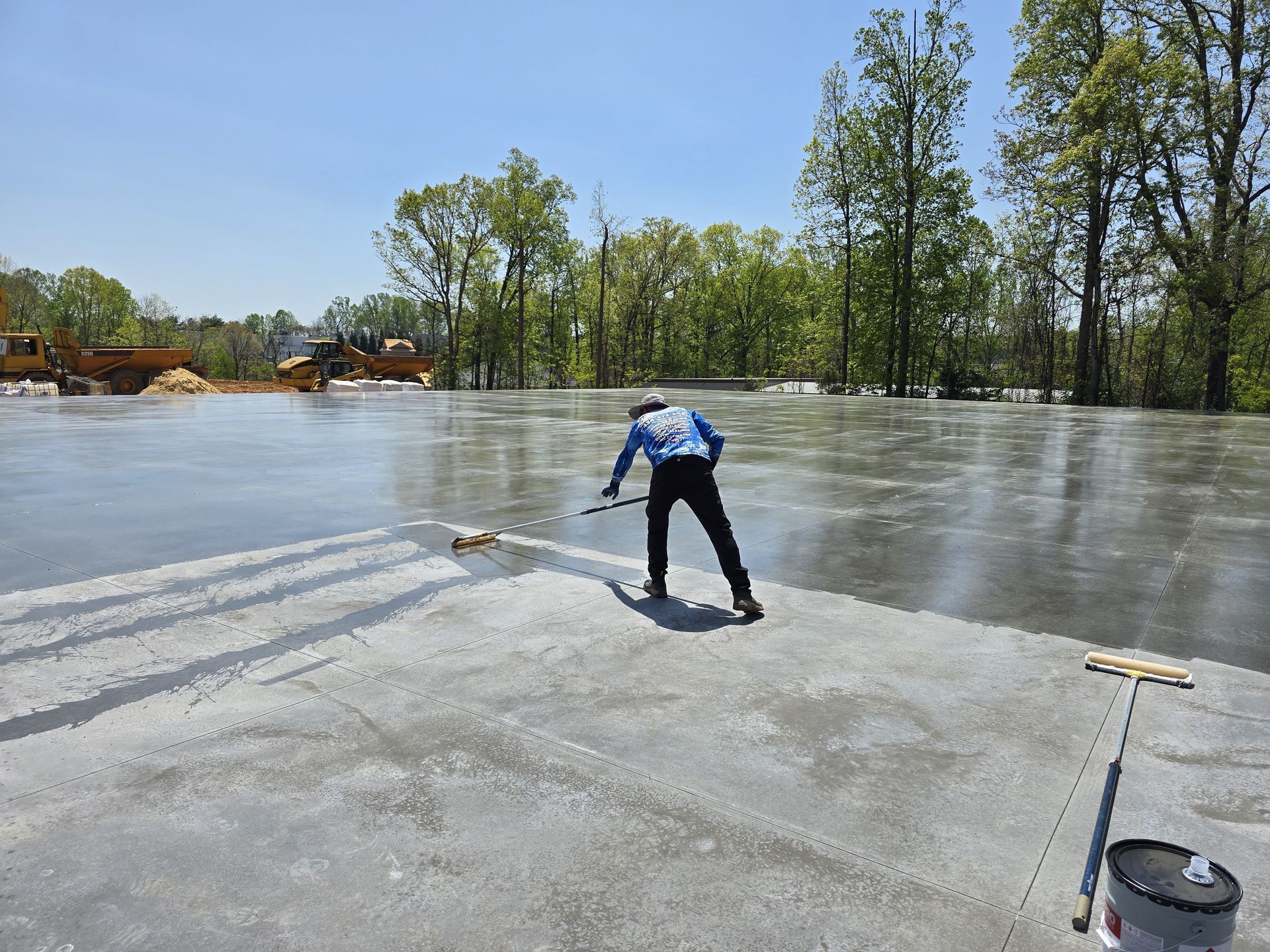 Person smoothing wet concrete with a long-handled tool on a sunny day. Construction site with trees in the background.