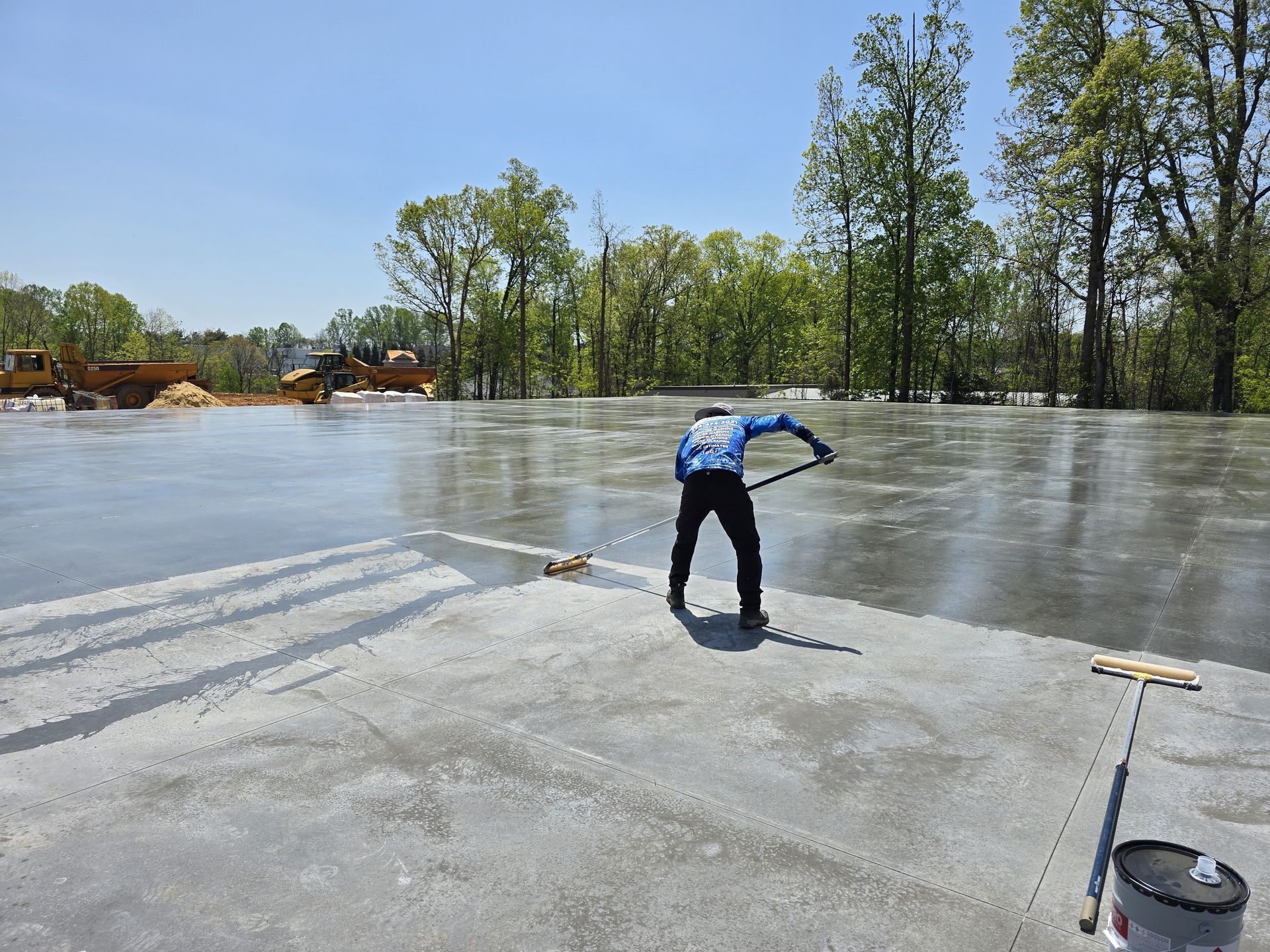 Worker smoothing wet concrete floor with a long-handled tool on a sunny day.