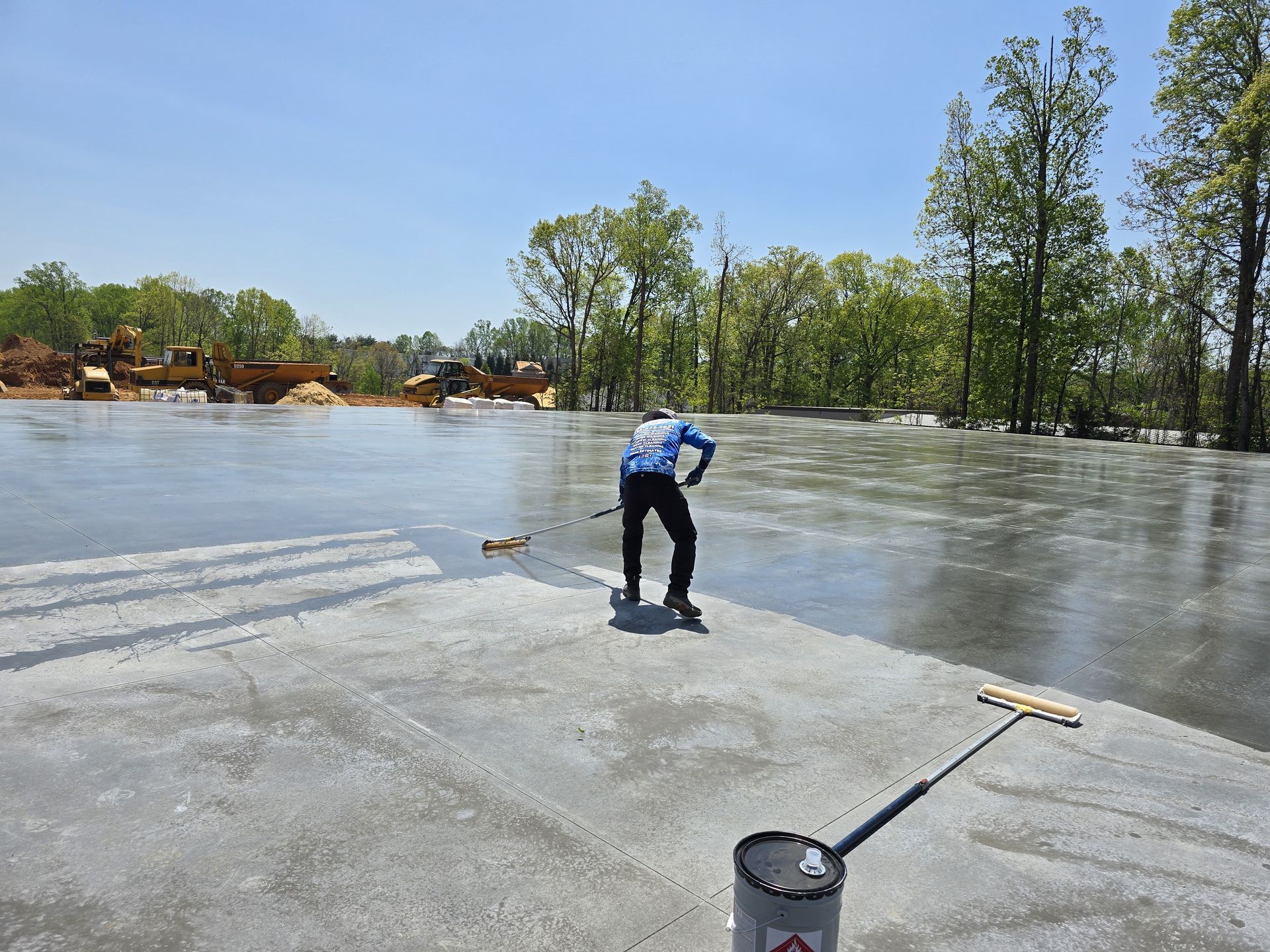 Person smoothing wet concrete on a large, open surface with tools, trees and sky visible.