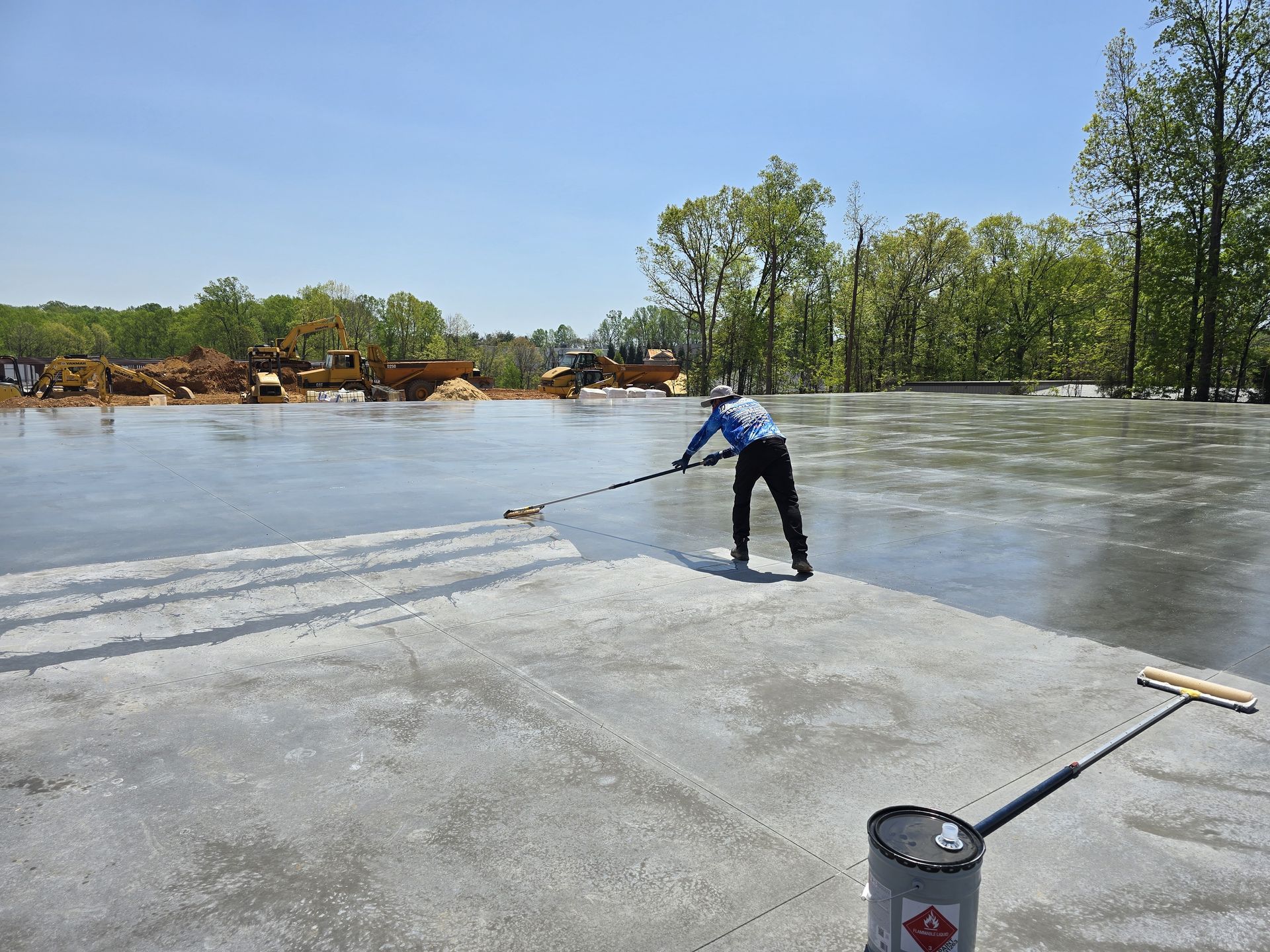 Worker smoothing wet concrete floor with a long-handled tool on a sunny day.