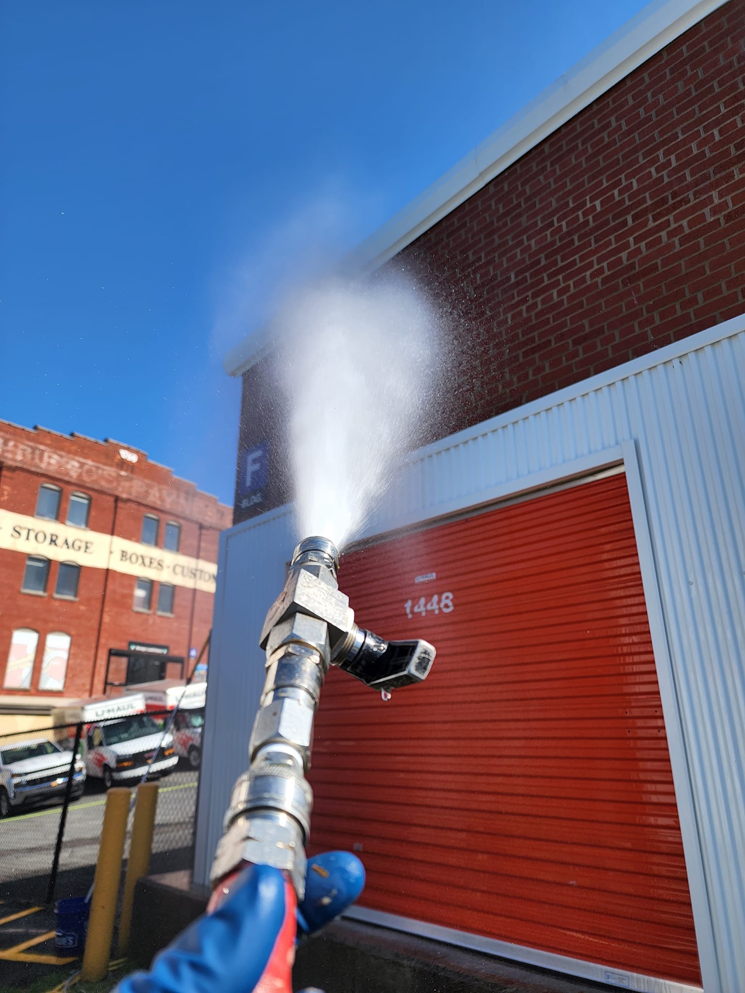 Person spraying a brick building with a high-pressure water gun. Blue sky. Red door and adjacent white metal building.
