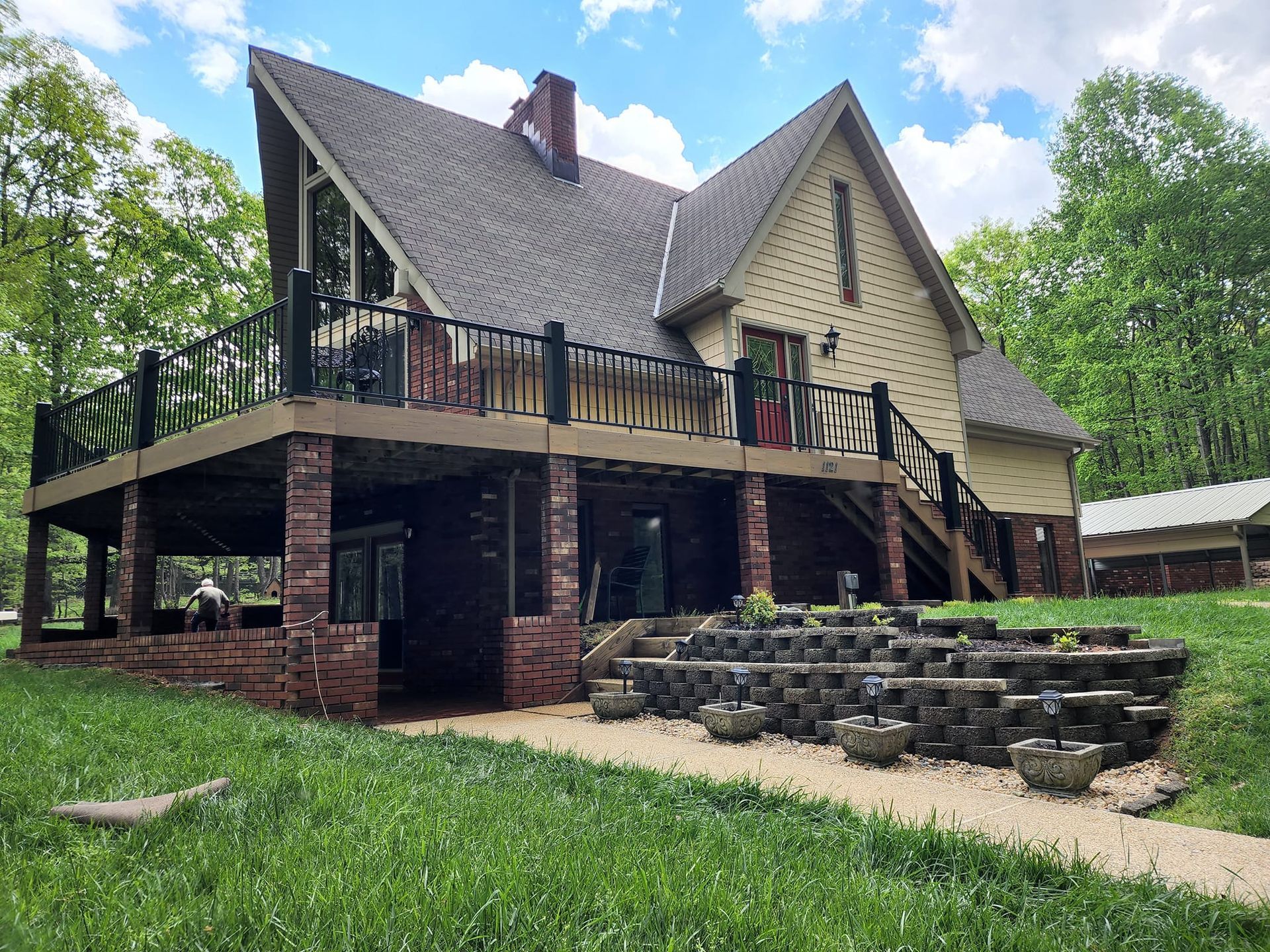 House with a large deck, brick columns, and multi-tiered planters in a grassy yard.