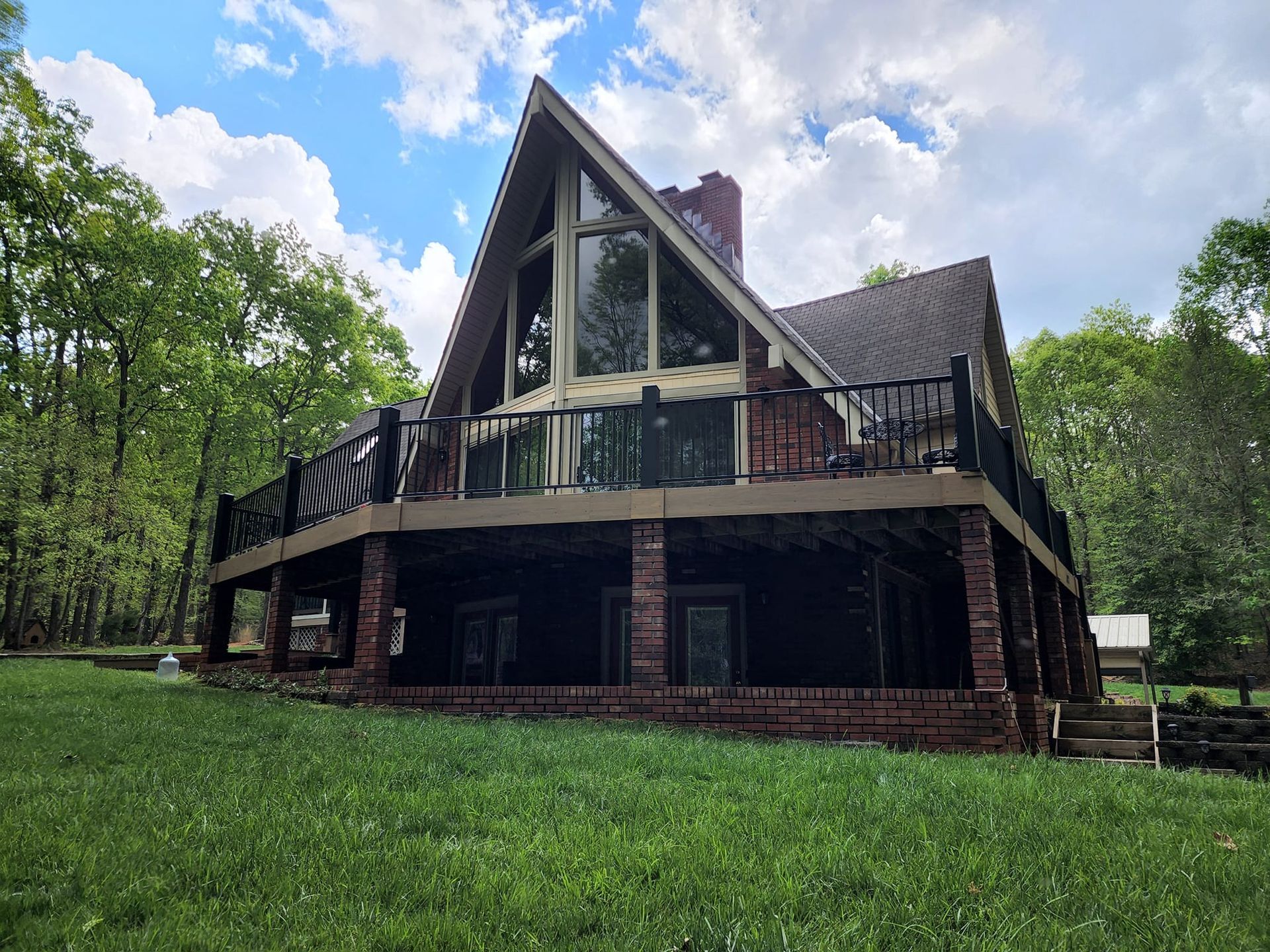A-frame house with a large deck and a brick base surrounded by trees and green grass.