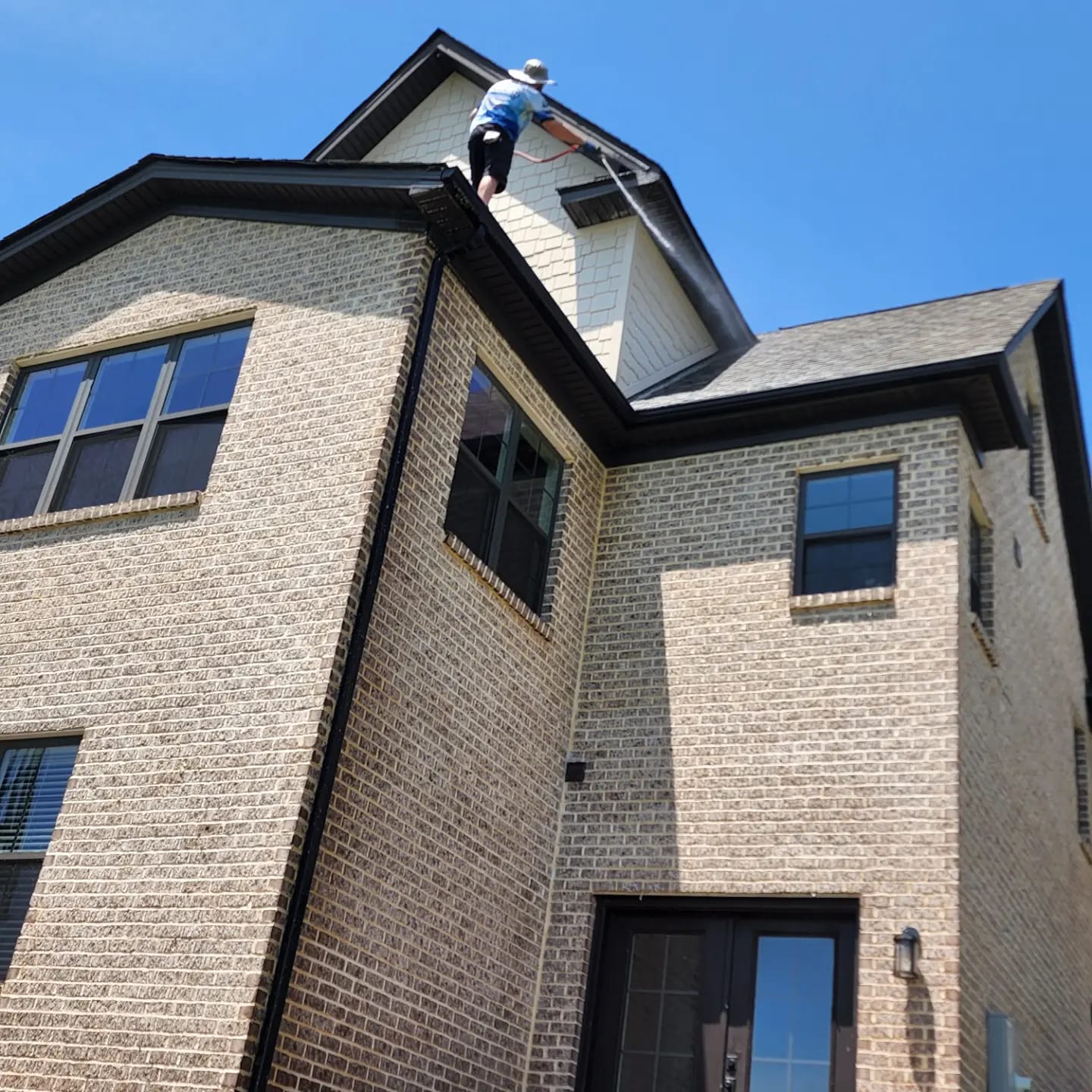 Man on a roof power washing a light brick home with black trim under a clear blue sky.