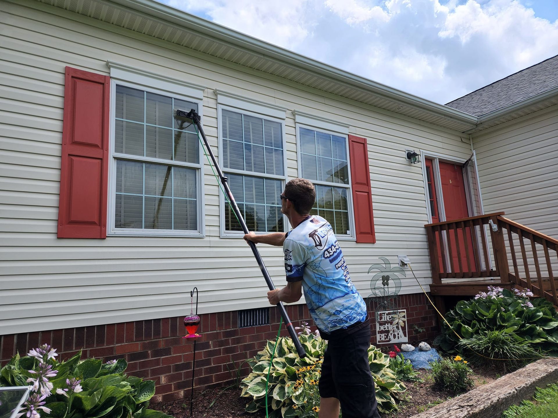 Man cleaning windows with a long-handled tool on a white house with red shutters.