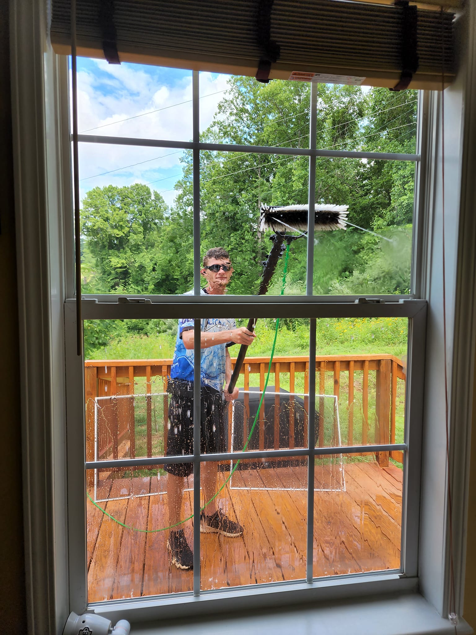 Man cleaning a window from a deck. Bright daylight, green trees in background.