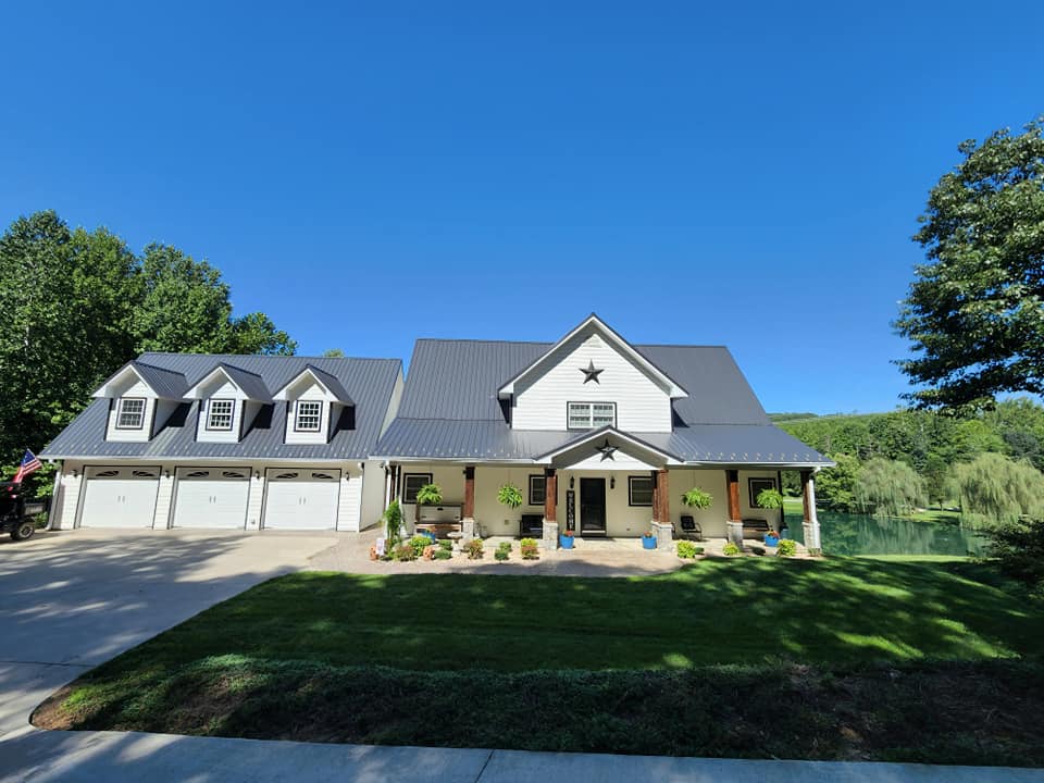 White farmhouse with a black roof, surrounded by green grass and trees, under a bright blue sky.