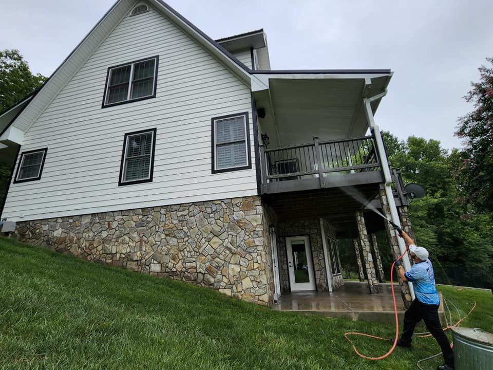 Person pressure washing a white house with stone foundation and a deck.