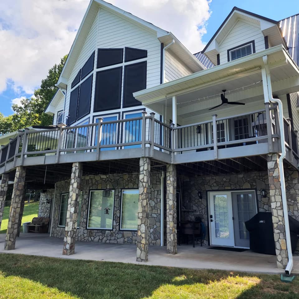 Two-story white house with a stone foundation and wrap-around deck; blue sky backdrop.