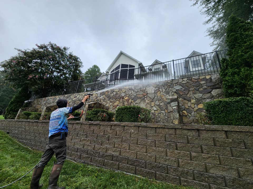 Person pressure washing a stone retaining wall, spraying water towards a house. Overcast day.