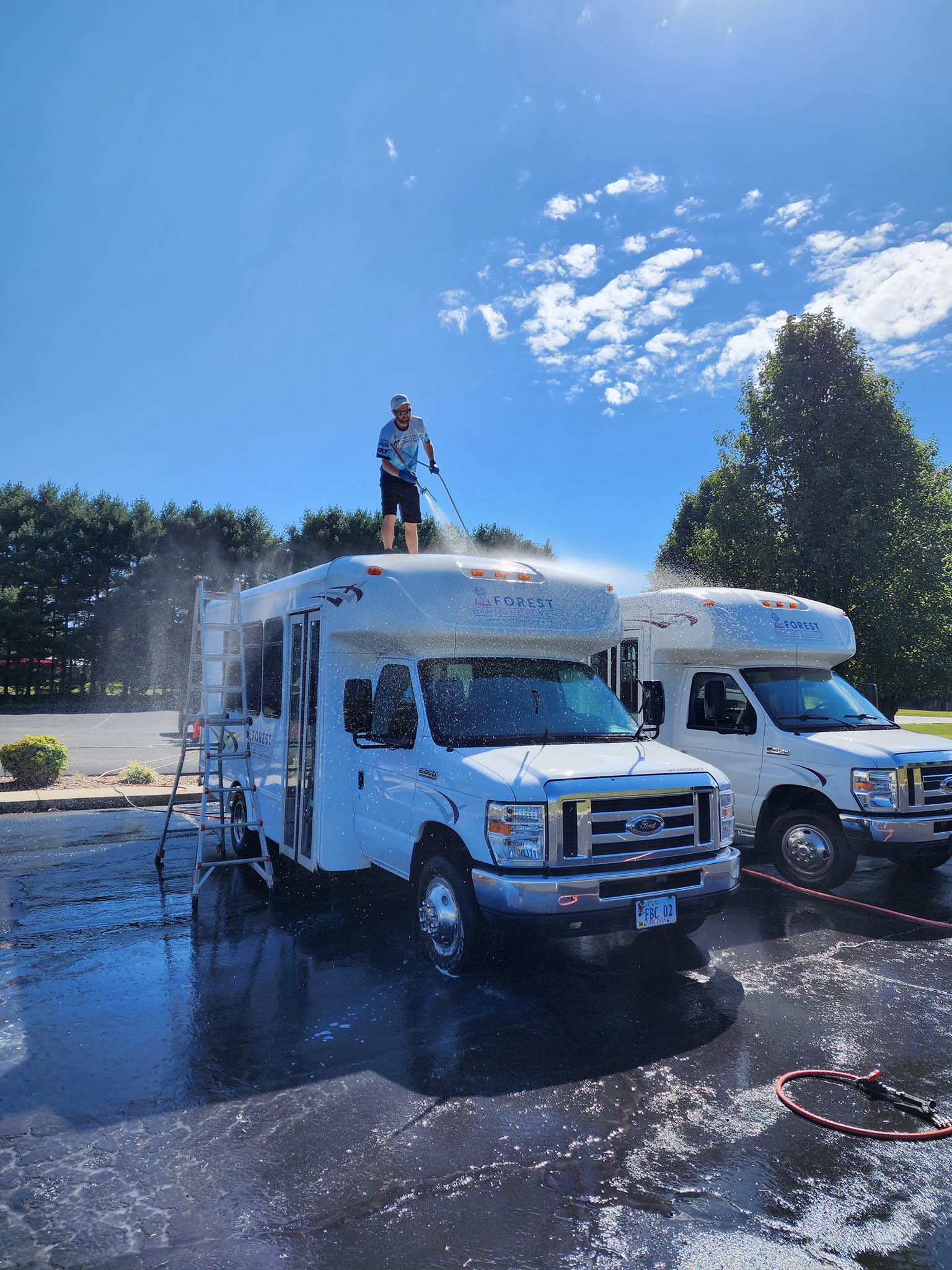 Person washing RV roof with a hose on a sunny day. Two white RVs parked outdoors.