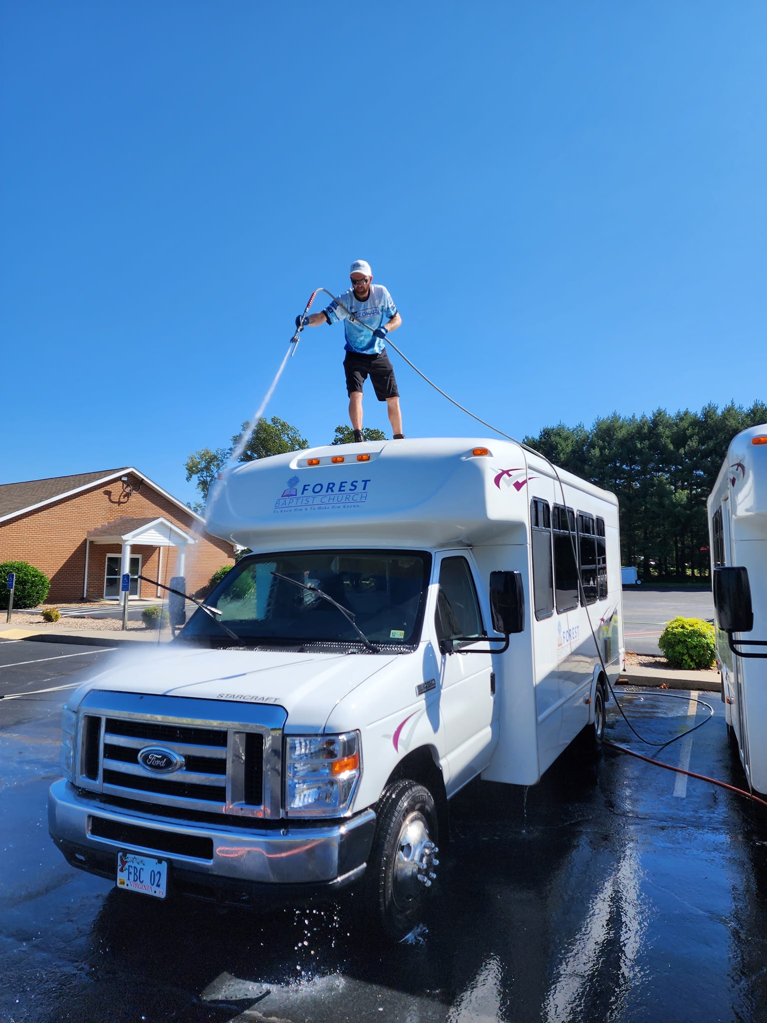 Man washing a white bus from its roof on a sunny day.