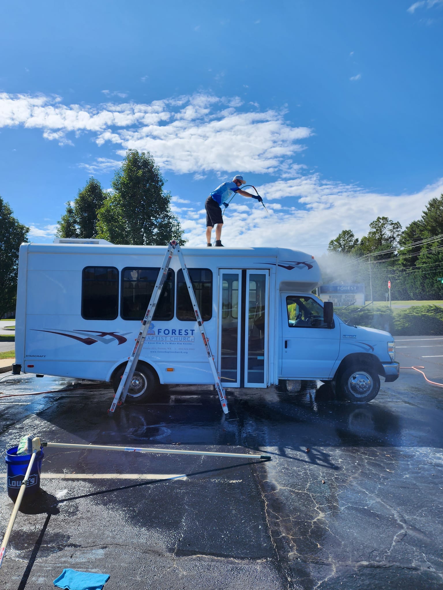 Man washing white bus roof with pressure washer, on a ladder, blue sky.
