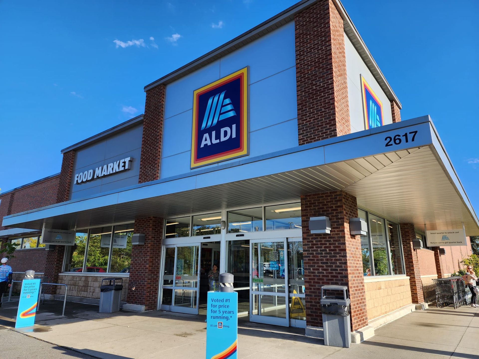 ALDI grocery store exterior, brick facade with blue ALDI logo. Sunny day, entrance visible, address 2617.