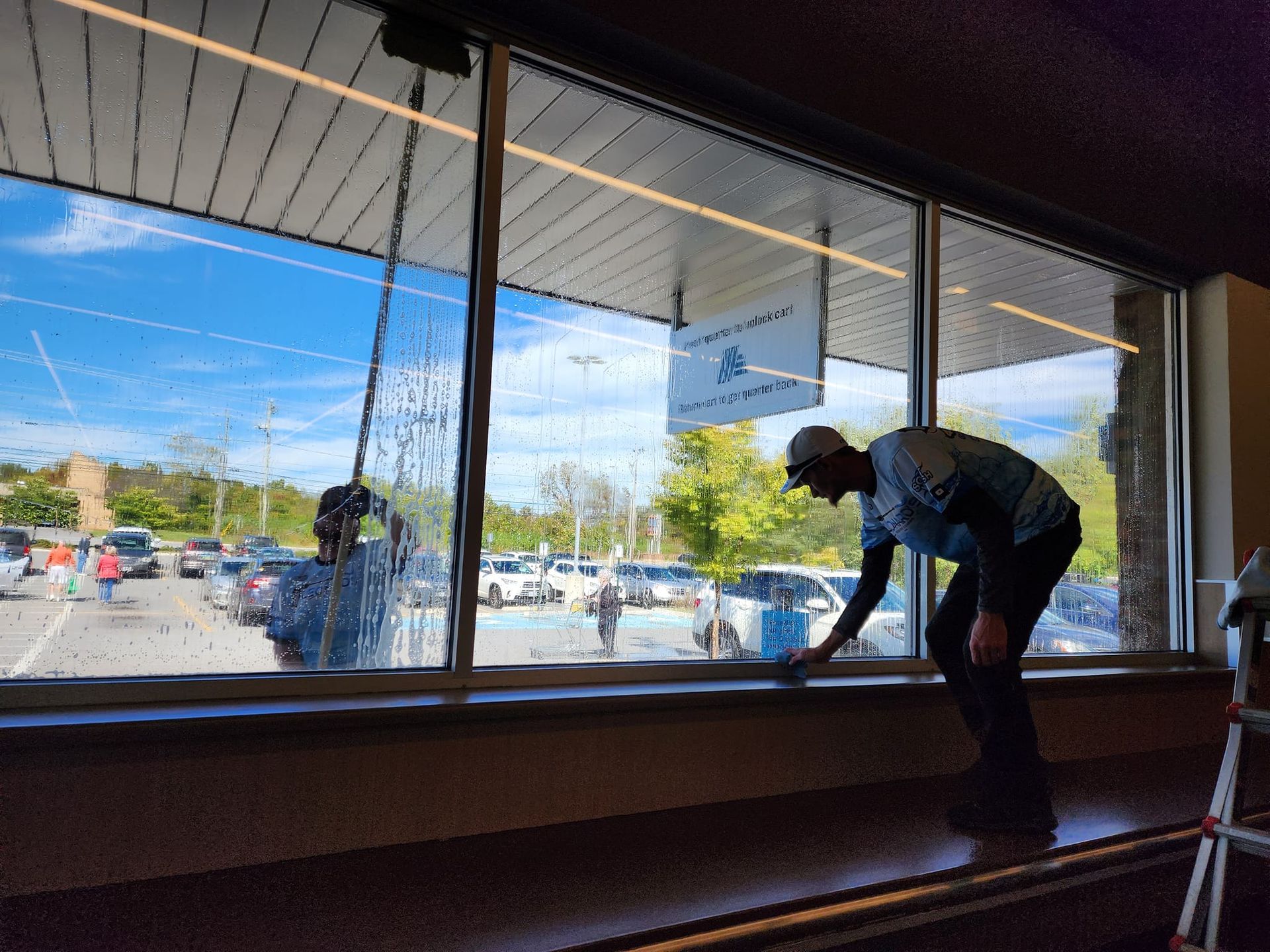 Man cleaning a large window, overlooking a parking lot, using a squeegee on a bright day.