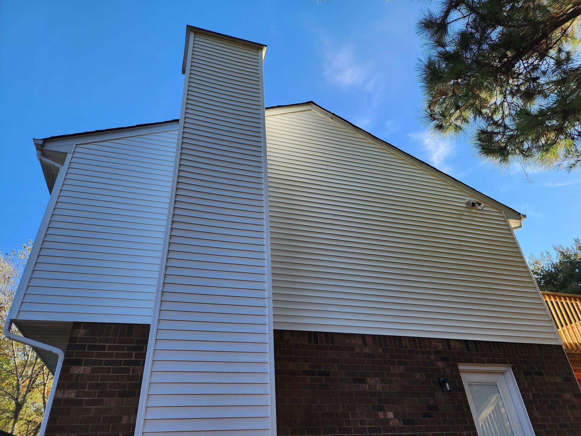 Side view of a two-story house with white siding, a brick base, and a tall chimney against a blue sky.
