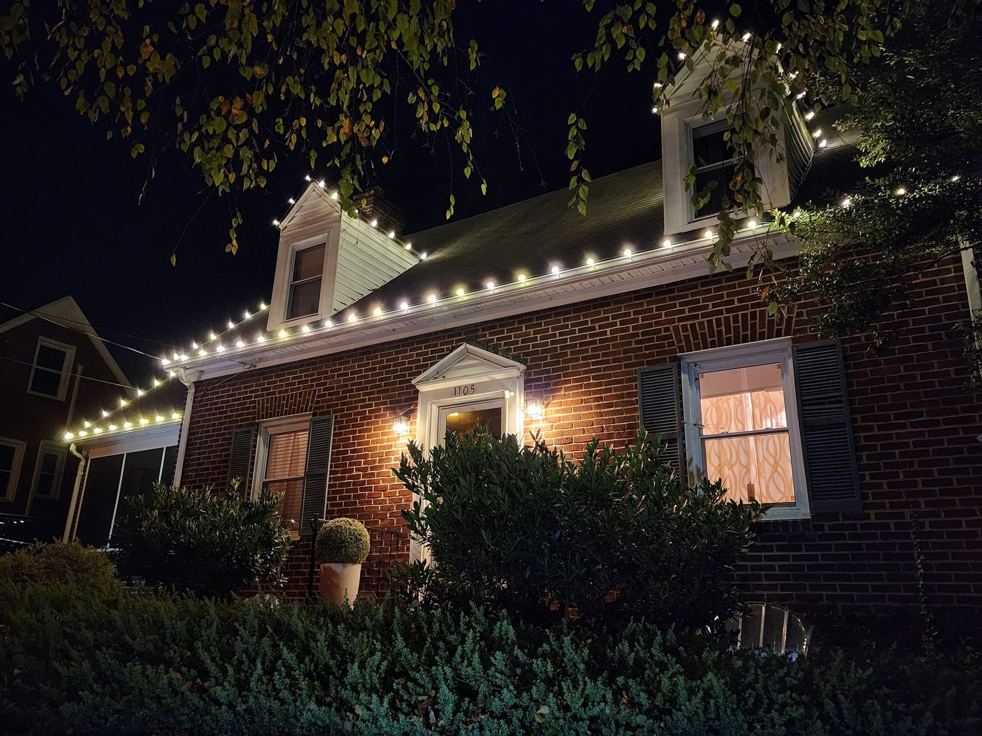 Brick house at night, lit with white lights on the roof. Bushes and a small tree in front.