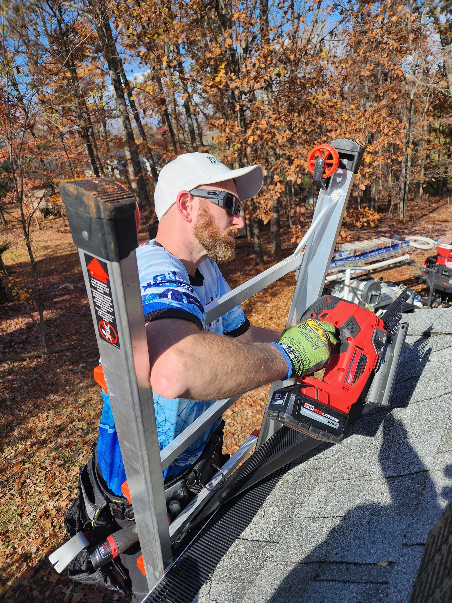 Man on a roof using a nail gun, wearing a hat and sunglasses. Fall foliage in background.