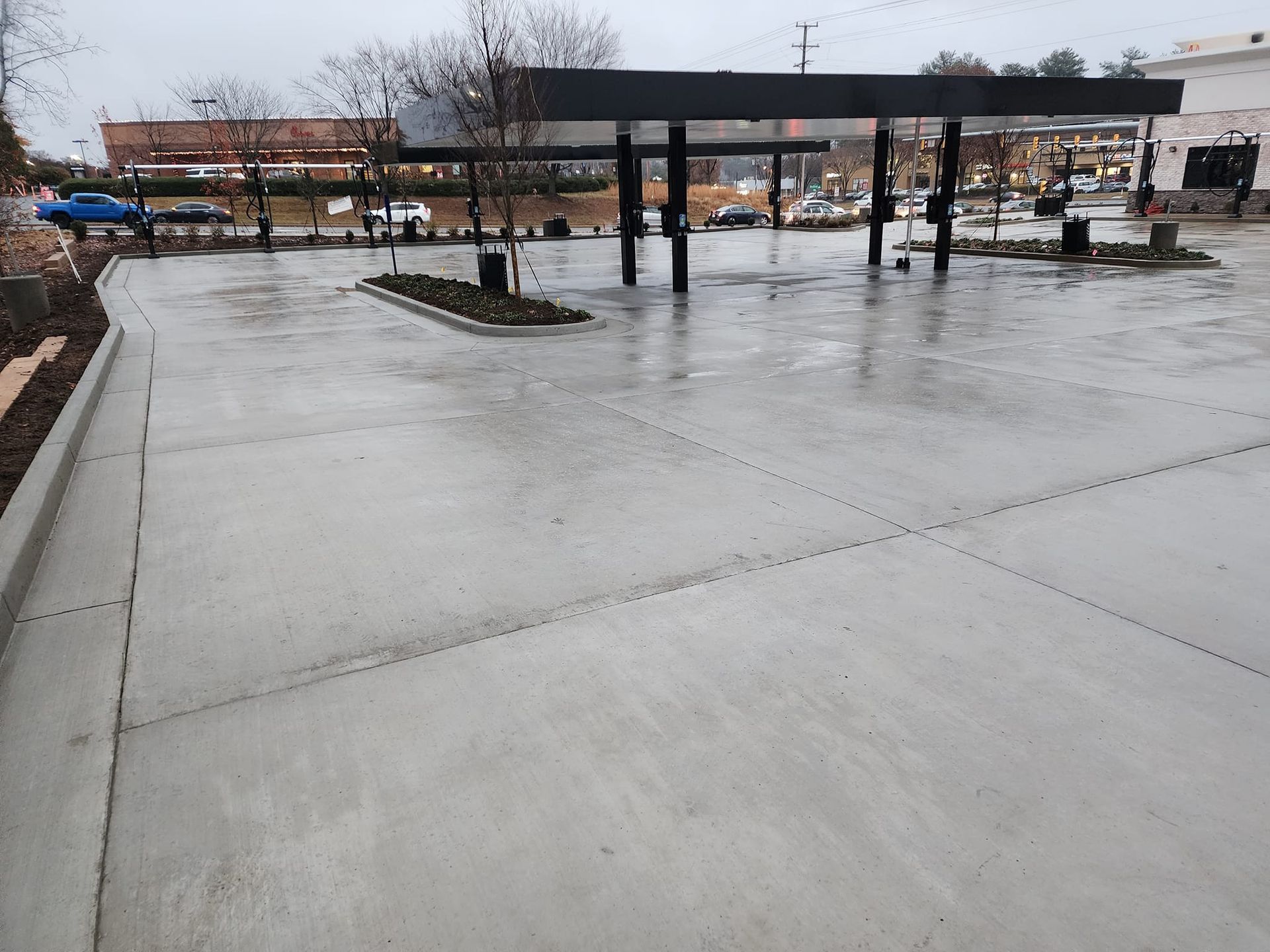 Wet, gray concrete at a gas station canopy with support pillars and a small island with plants.