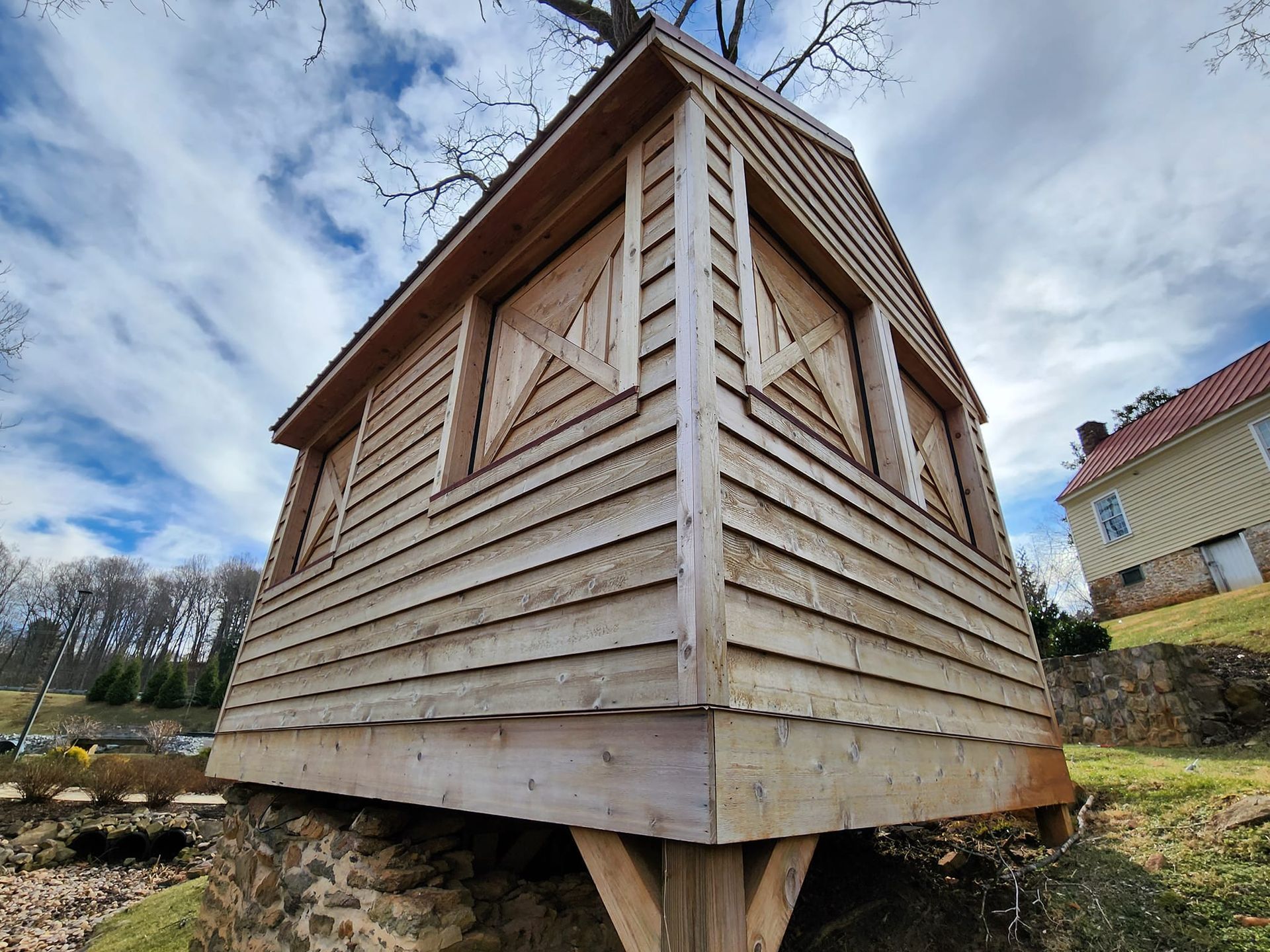 Wooden shed-like structure with angled walls, supported by posts, set on a hillside with a cloudy sky.