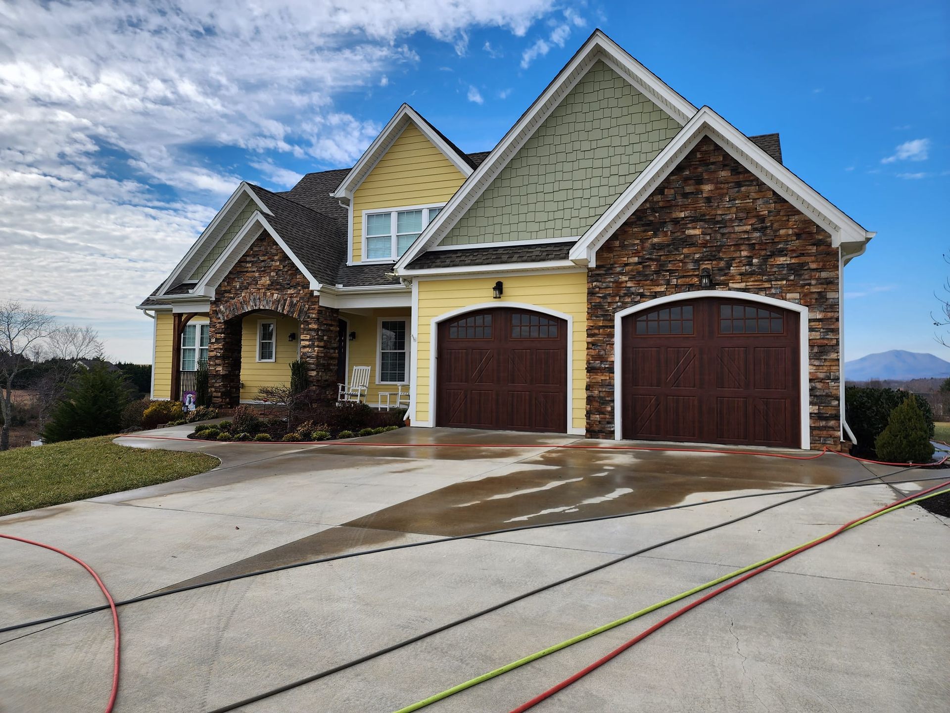 Two-story yellow house with brown garage doors and stone accents, on a wet driveway under a blue sky.