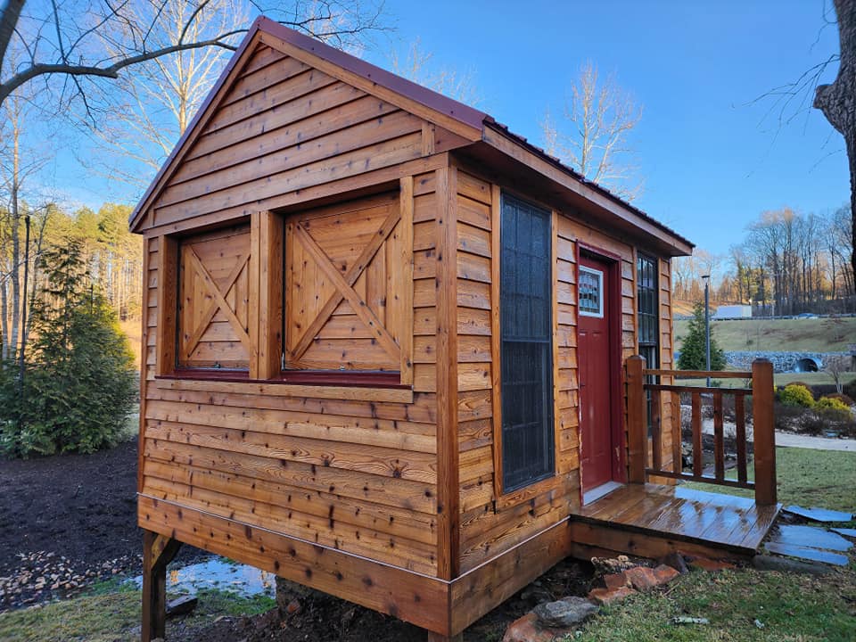 Small wooden shed with red door, shutters, and roof; set outdoors on a deck.