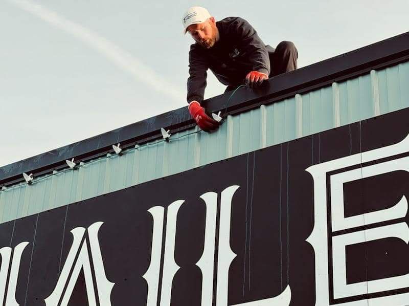 Man in a white hat and gloves installing a sign on a building rooftop.