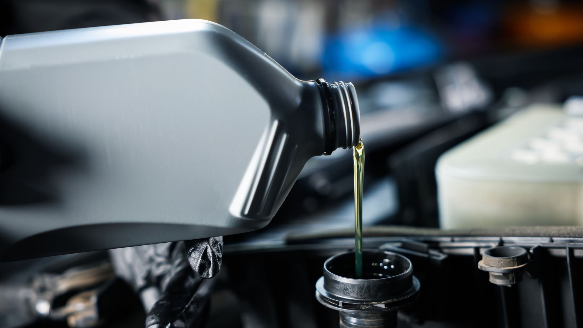 Motor oil pouring from a gray jug into a car's engine.