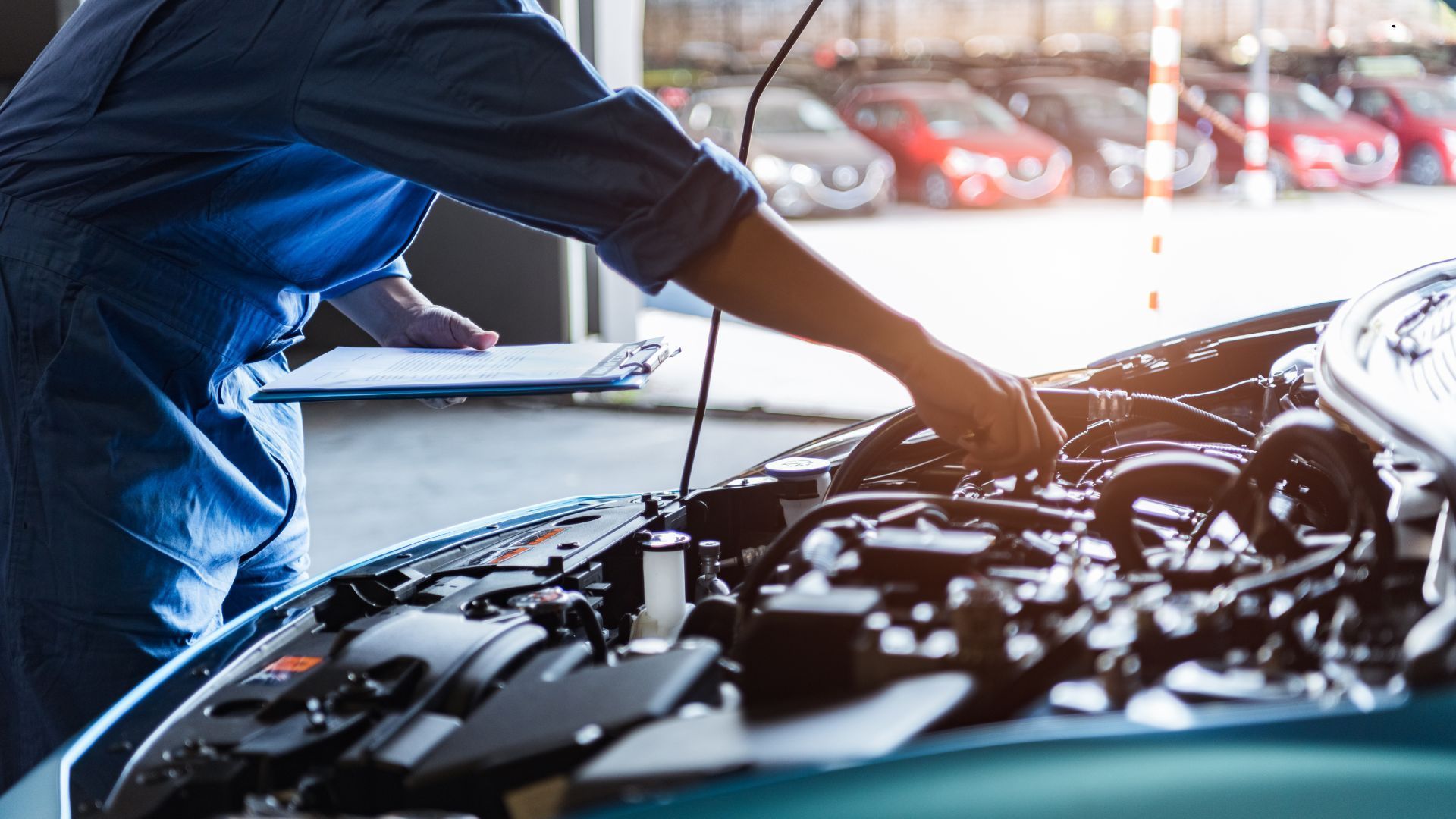 Mechanic in blue jumpsuit checks car engine with clipboard in an auto shop.