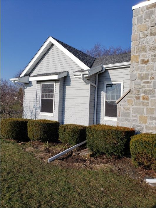 Light gray house with dark roof, windows, and stone chimney; green hedges in front.