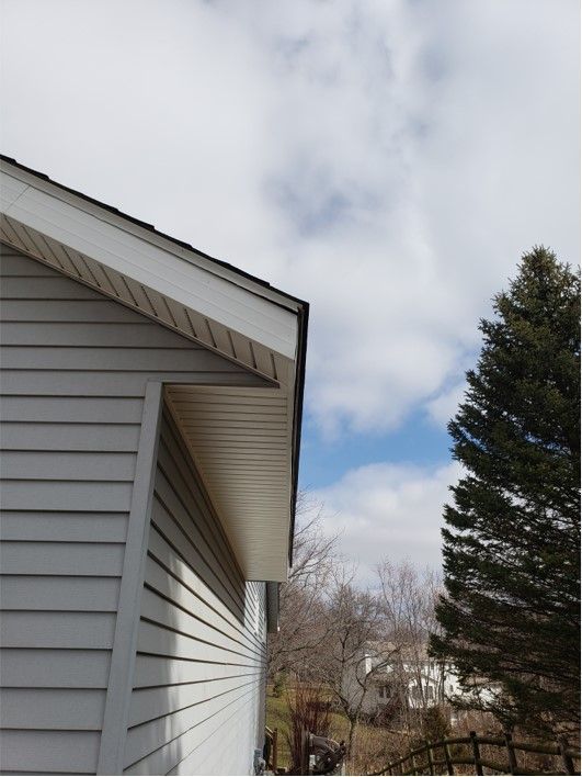 Gray siding on a house with a white soffit and blue sky with clouds. A tall evergreen tree is to the right.