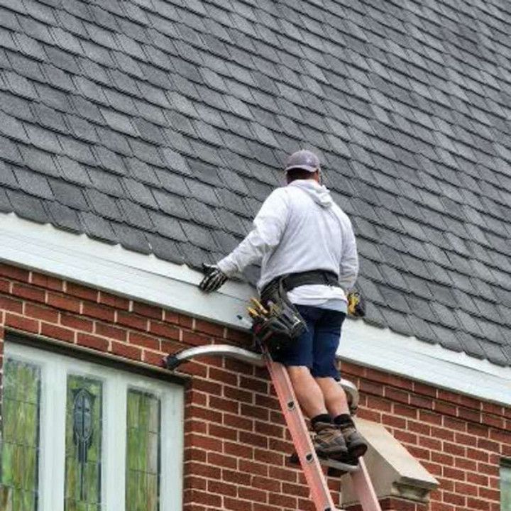 Roofer on a ladder inspecting a gray shingled roof, brick wall, and white trim.