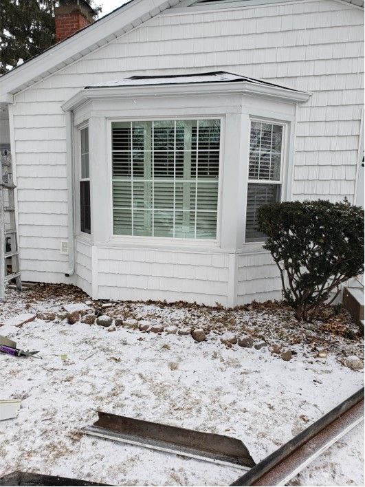 White house with bay window and snow-covered ground, small shrub on right.