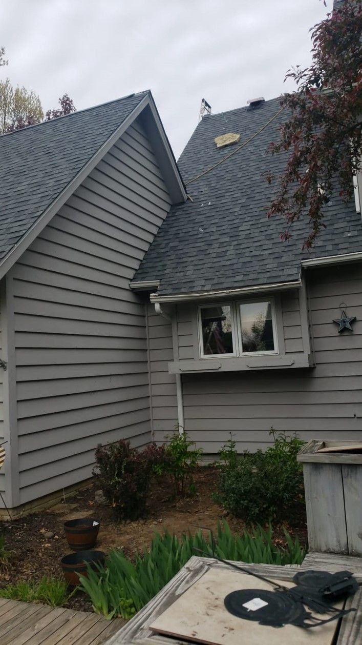 Gray house exterior with multiple roof sections and a small window. Shrubs and a wooden deck are visible.