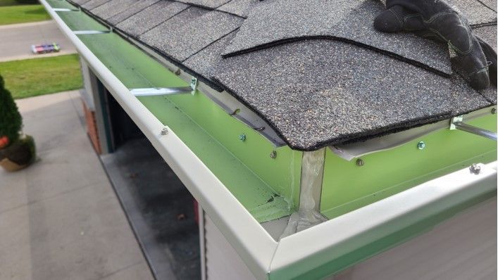Green gutter installed along a gray shingled roof. White trim is visible on the side.