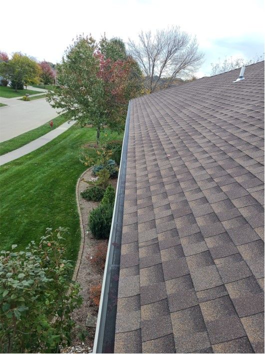 Brown shingled roof with a gutter and green yard with trees and a sidewalk in the background.