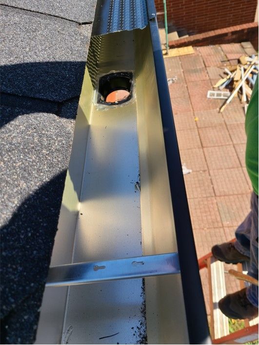 Gutter with debris, screen, and downspout opening. A person is on a ladder.