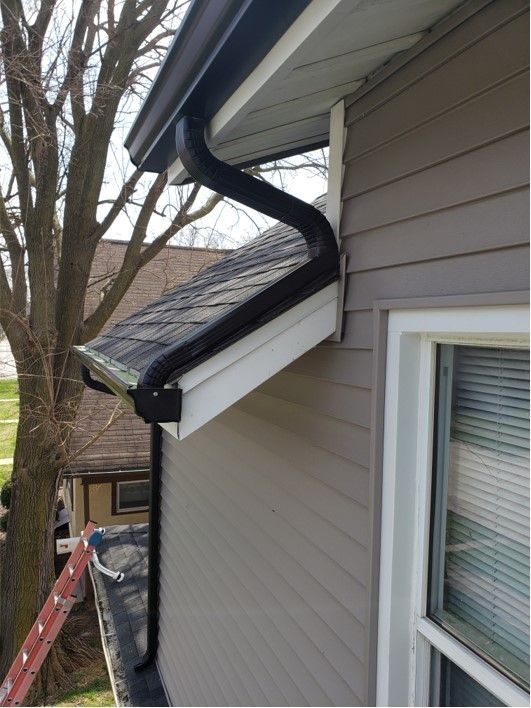 Black rain gutter on a house with tan siding, downspout curves downward.