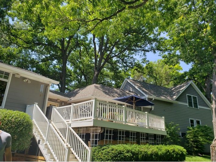 A two-story house with a large deck, surrounded by trees and bushes under a blue sky.