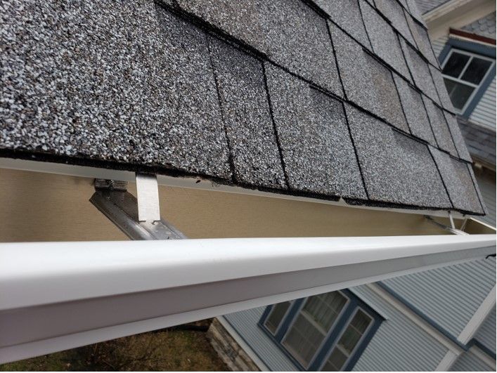 White gutter under a gray shingled roof on a house. Supports are visible.