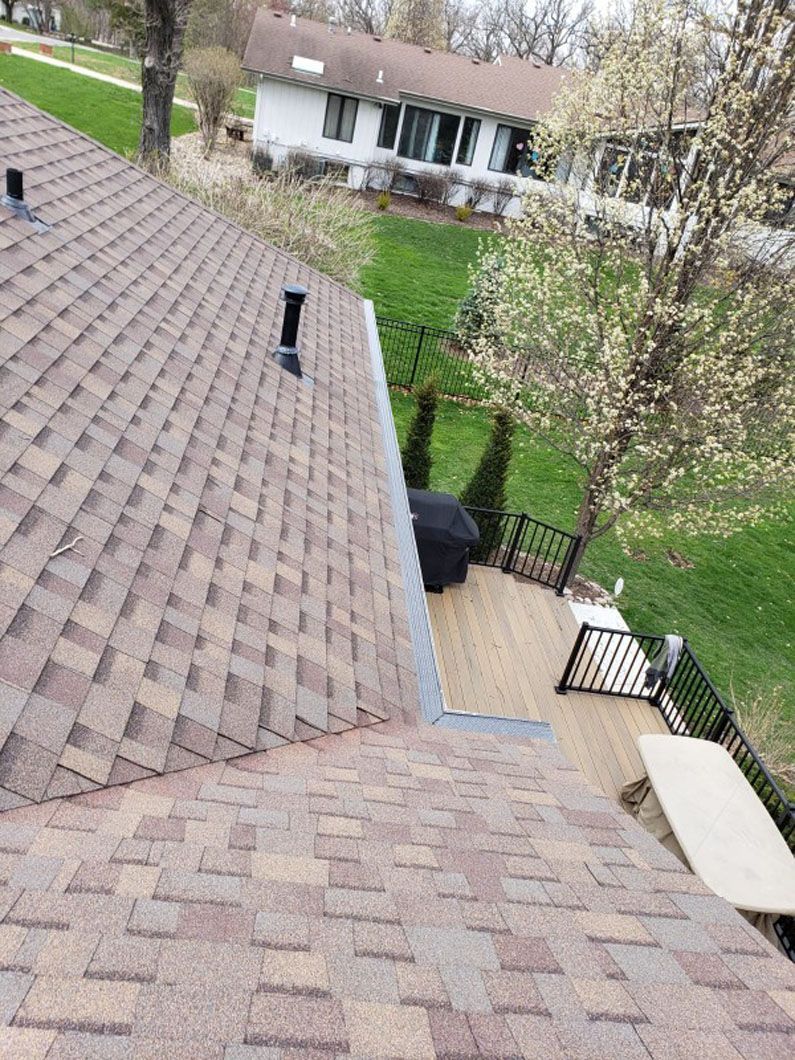 Shingled roof with a black vent, overlooking a wooden deck and green lawn with a distant white house.