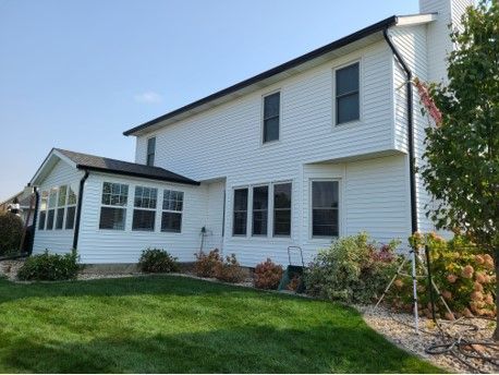 White house with black trim, windows, and gutters. Green lawn, landscaping, and blue sky.