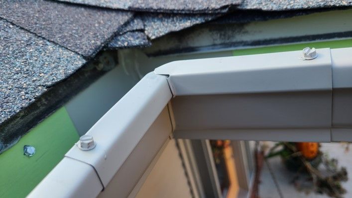 White gutter corner attached to a gray shingled roof, bolted to a green surface.