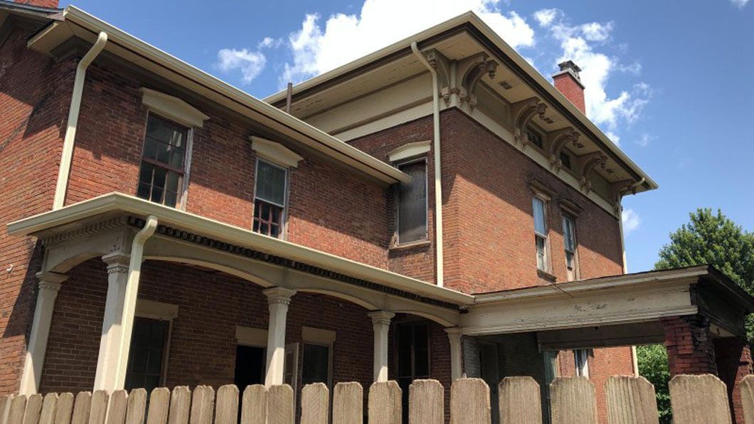 Two-story brick house with white trim, porch, and picket fence under a blue sky with clouds.