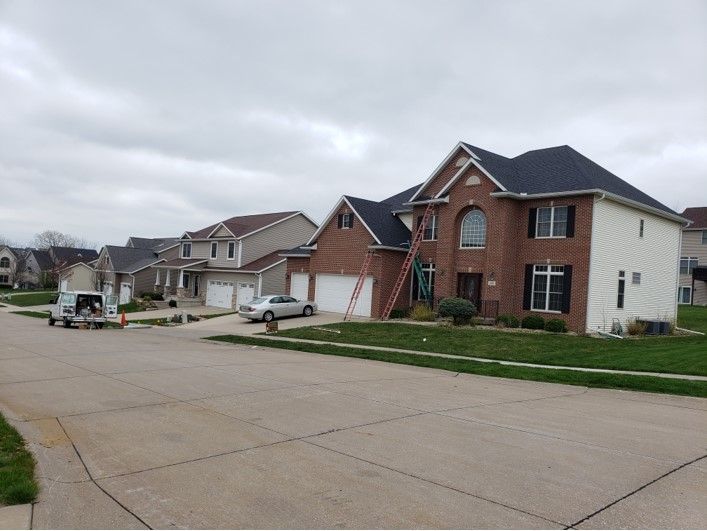 Suburban street with brick and siding houses. A ladder leans against a house, workers near. Gray sky.