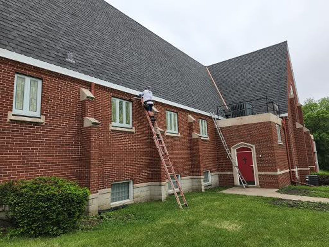 Person on a ladder next to a brick building with a dark gray roof and a red door.