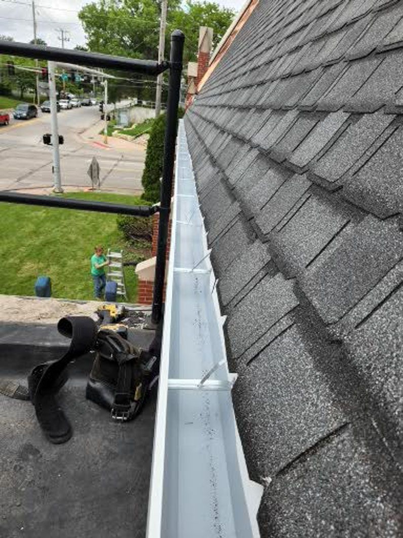 New gutter installed on a residential roof. Black asphalt shingles, scaffolding, and a person working.